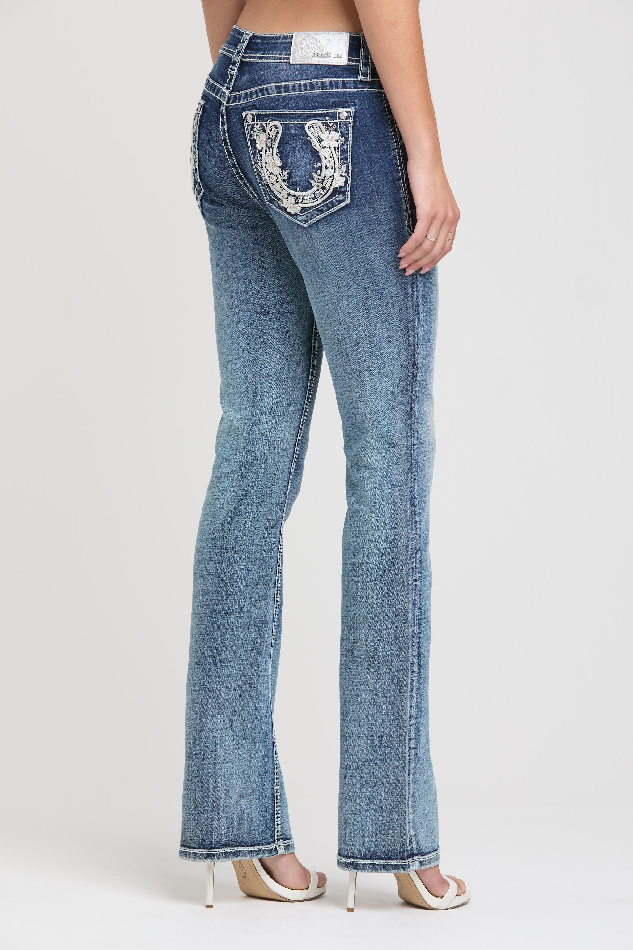 Grace in LA womens bootcut jeans in a medium featuring a side-rear view with silver horseshoe and white floral embroidery on the back pocket.