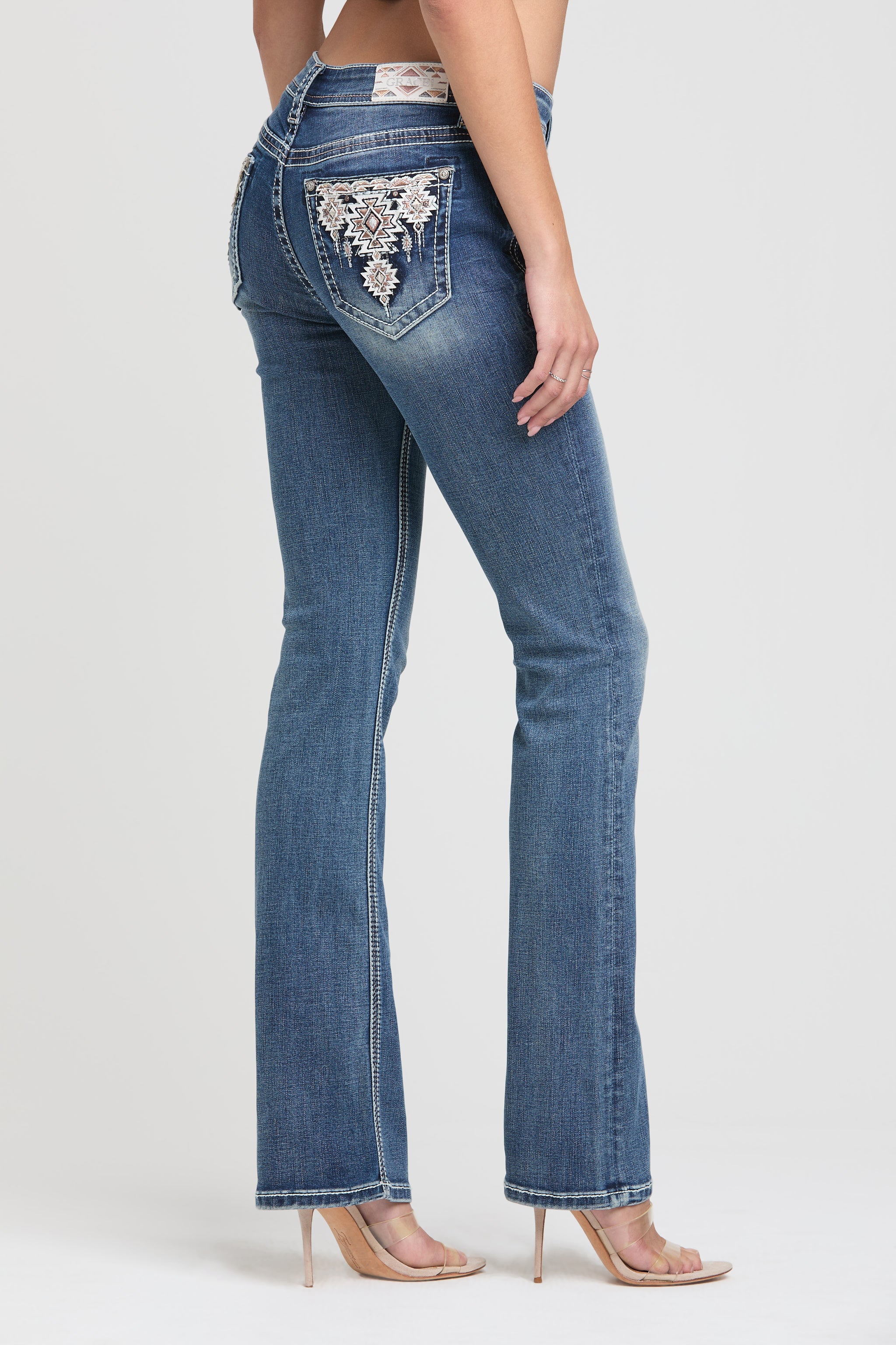 Grace in LA rhinestone jeans for women featuring a dark wash with a desert-themed brand patch and colorful geometric tribal embroidery on the back pocket.