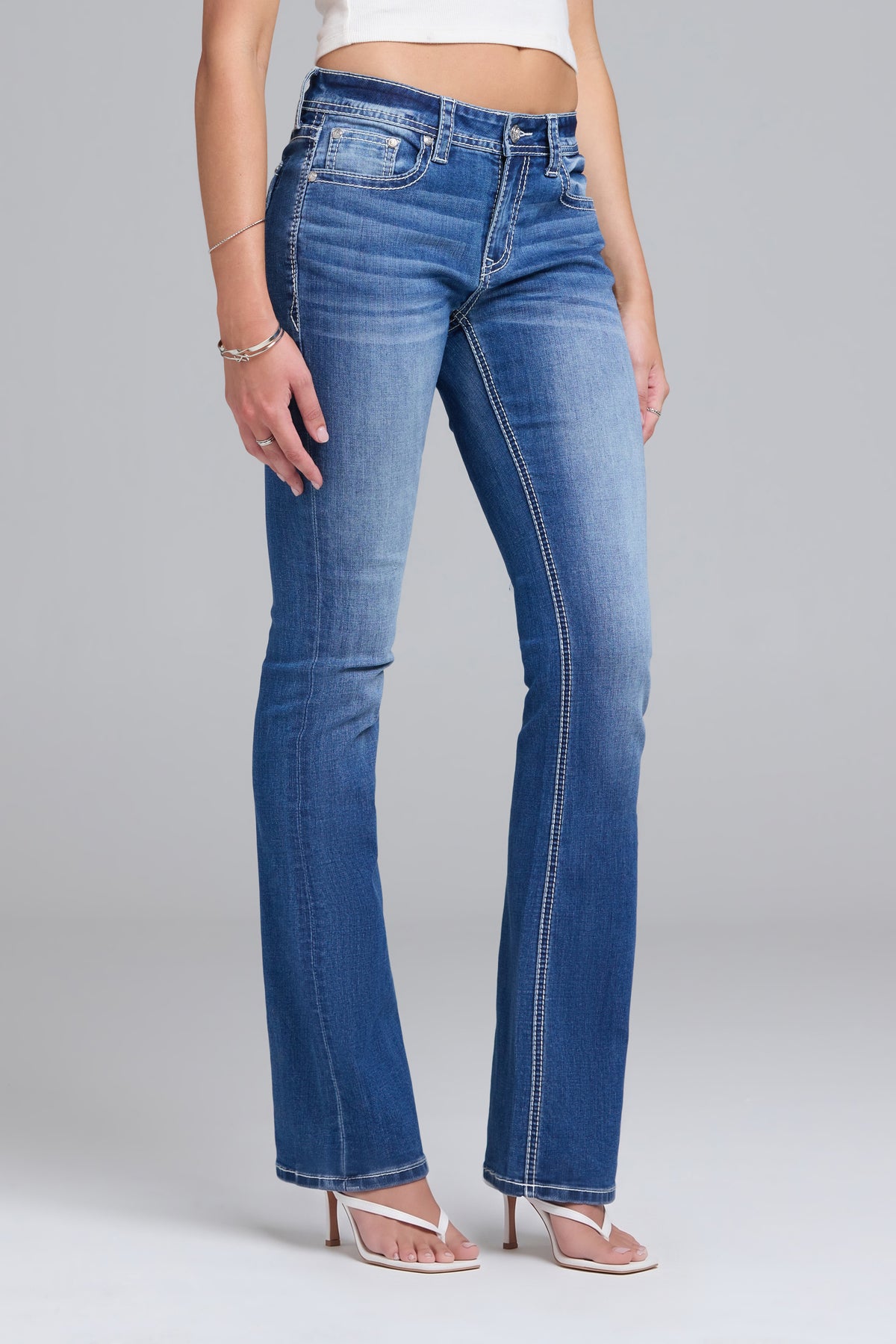 A model wearing a pair of medium-wash blue denim bootcut jeans with contrast stitching, paired with a white crop top and beige heeled sandals, against a light gray studio background.