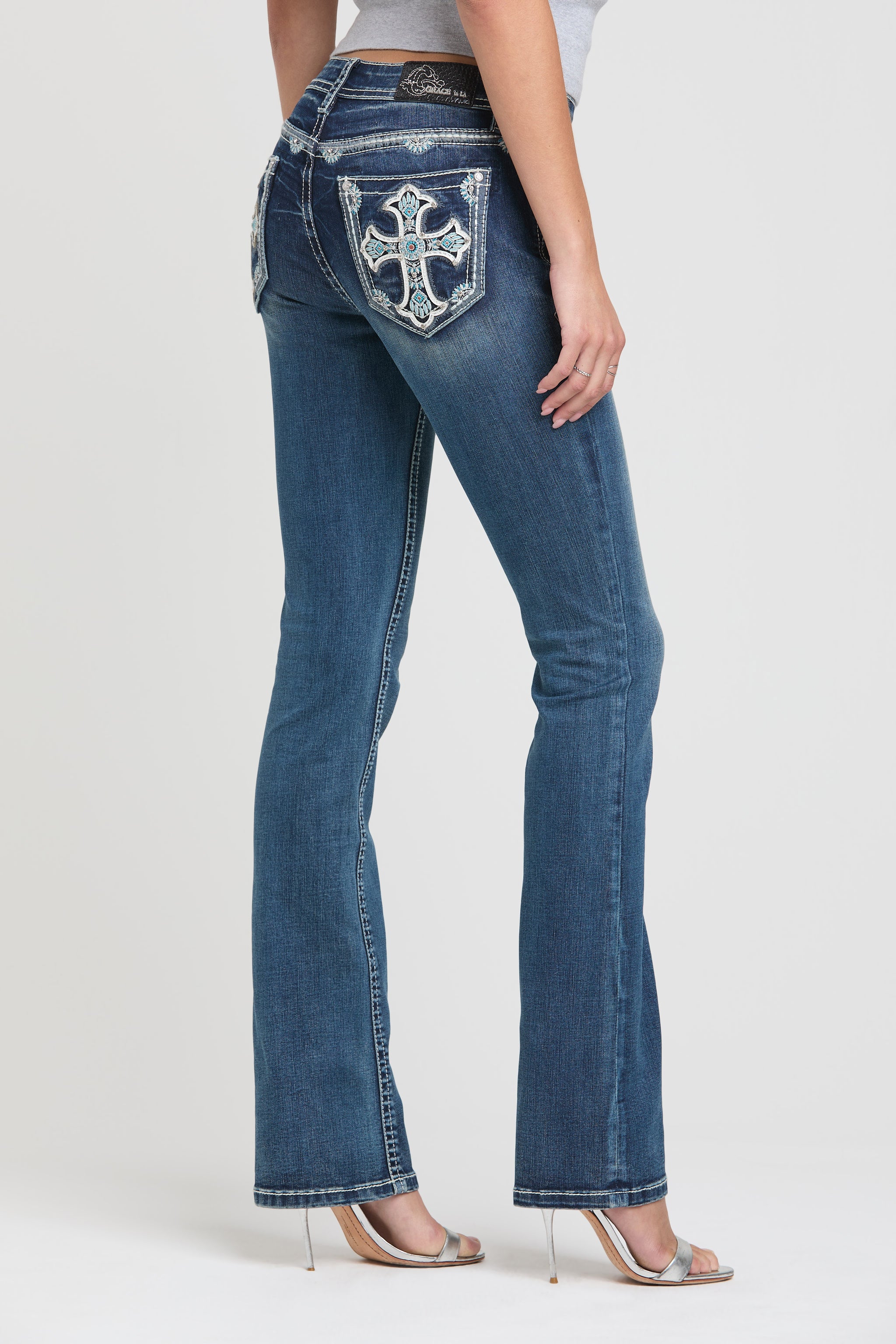 Grace in LA womens bootcut jeans in a dark wash featuring a full rear-view silhouette with ornate white embroidered crosses and turquoise accents on the pockets.