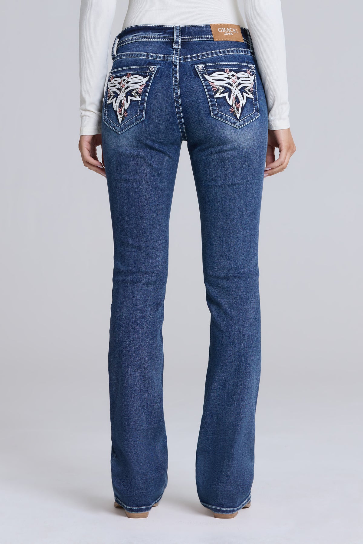 Rear view of a person wearing blue denim jeans with white embroidered winged motifs on the back pockets, paired with a white long-sleeve top, against a light gray studio background.