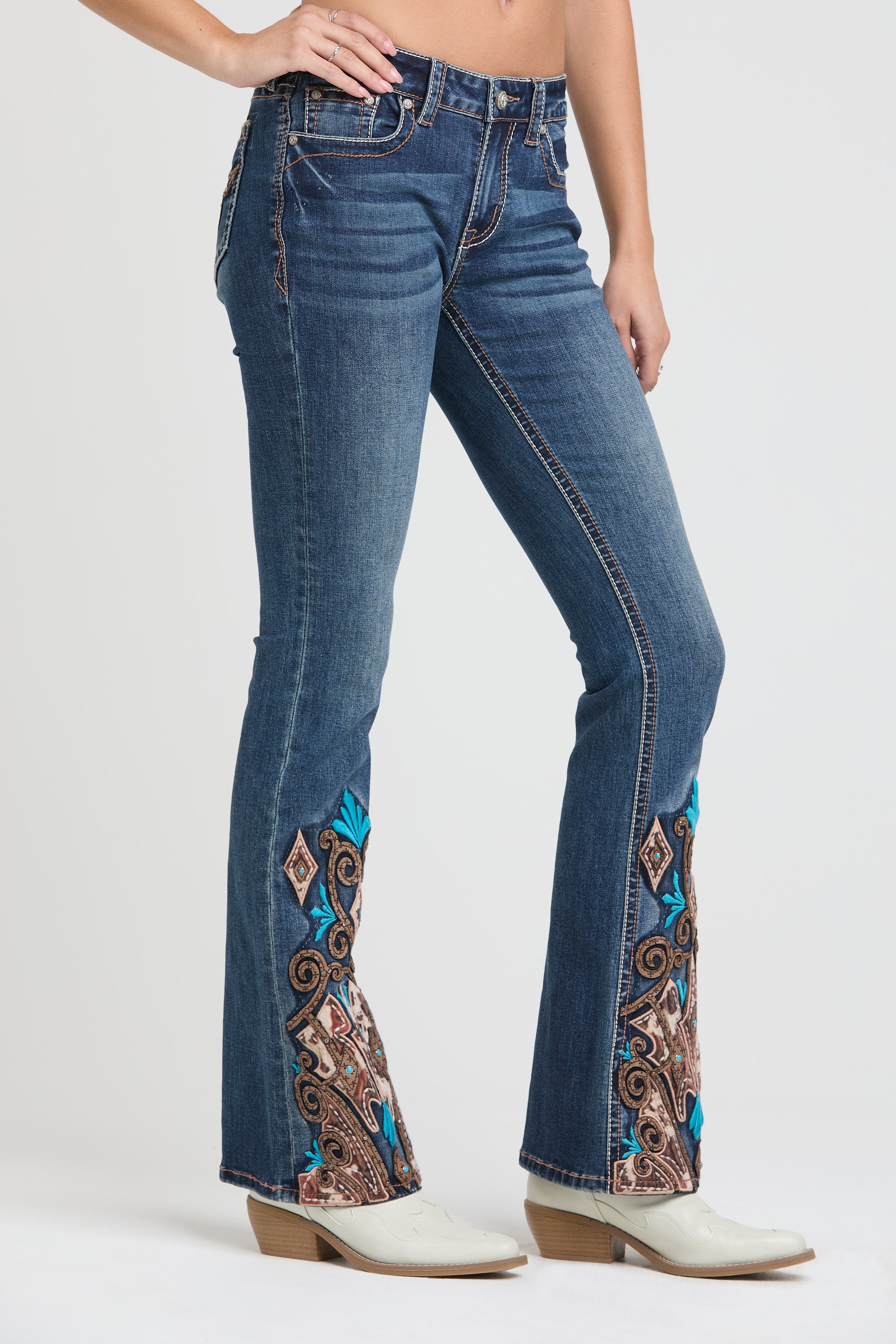 Grace in LA womens bootcut jeans in a dark featuring intricate western-inspired paisley and turquoise embroidery on the outer leg flare, styled with white cowgirl boots.