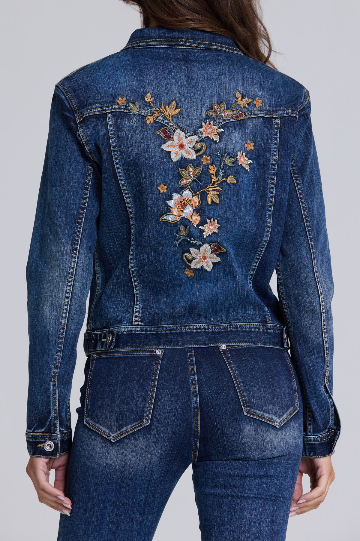 Back view of a model wearing a blue denim jacket with floral embroidery on the upper back and matching blue denim jeans, against a light gray studio background.