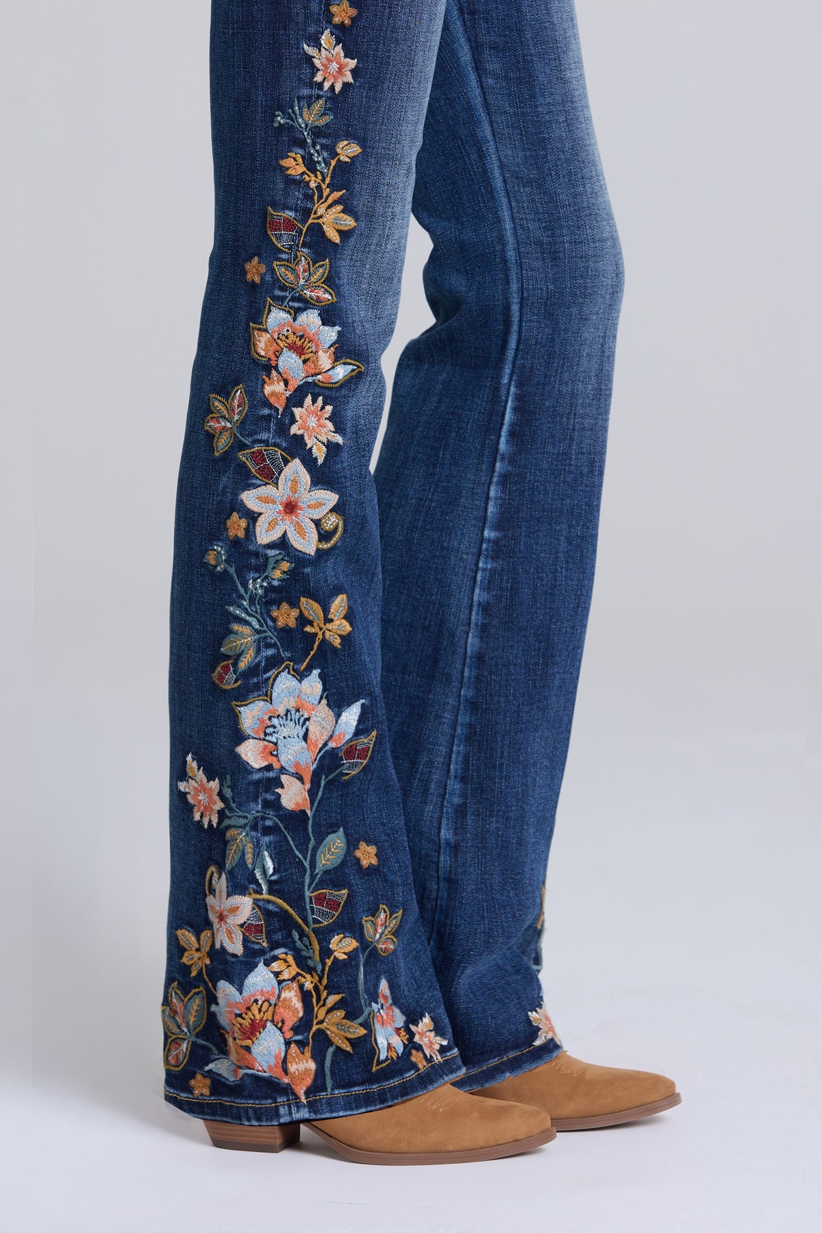 Close-up of a person wearing blue flare jeans with colorful floral embroidery down the outer legs, paired with tan suede boots against a light gray studio background.