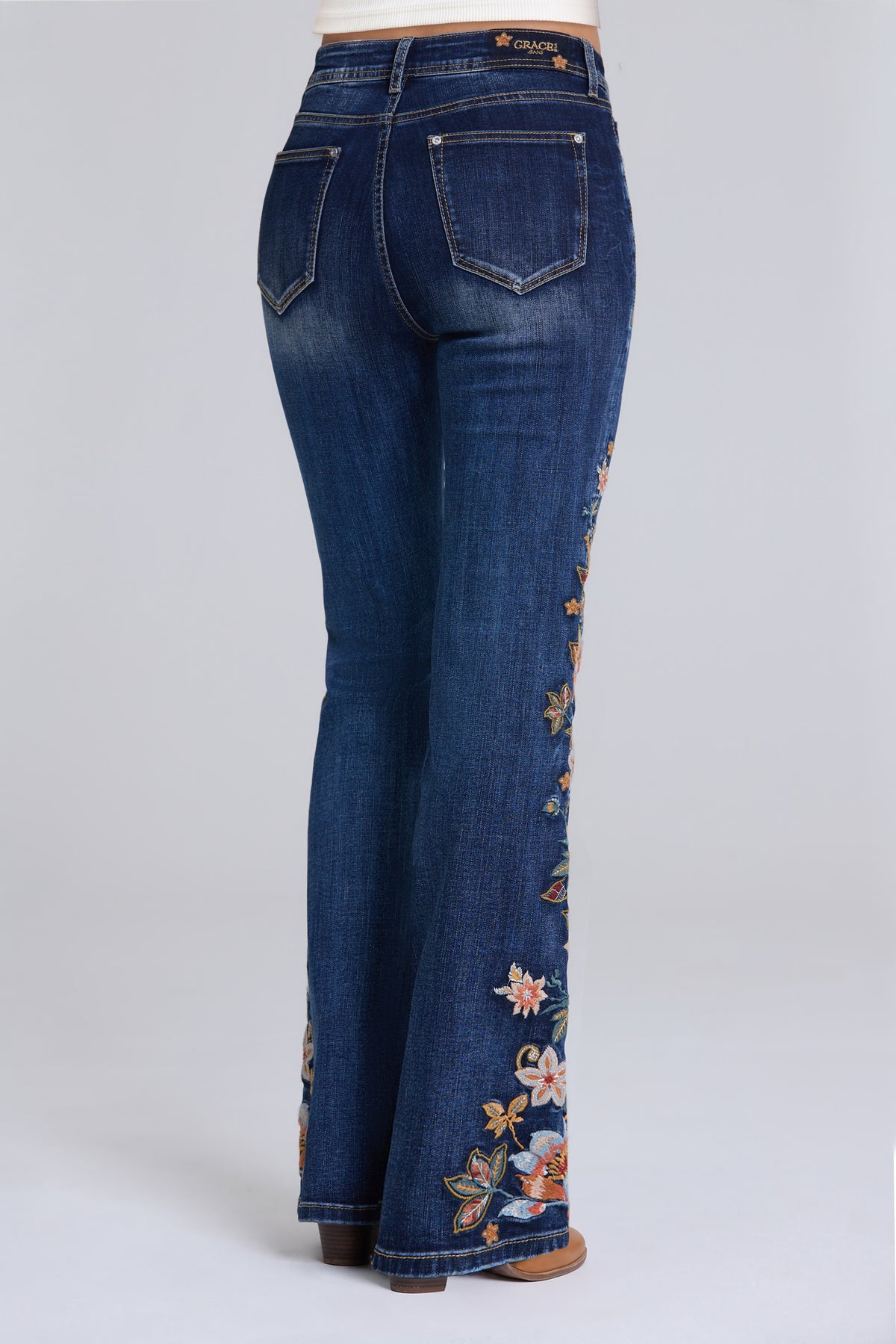 Rear view of a person wearing dark blue flare jeans with colorful floral embroidery down the outer leg, photographed against a light gray studio background.