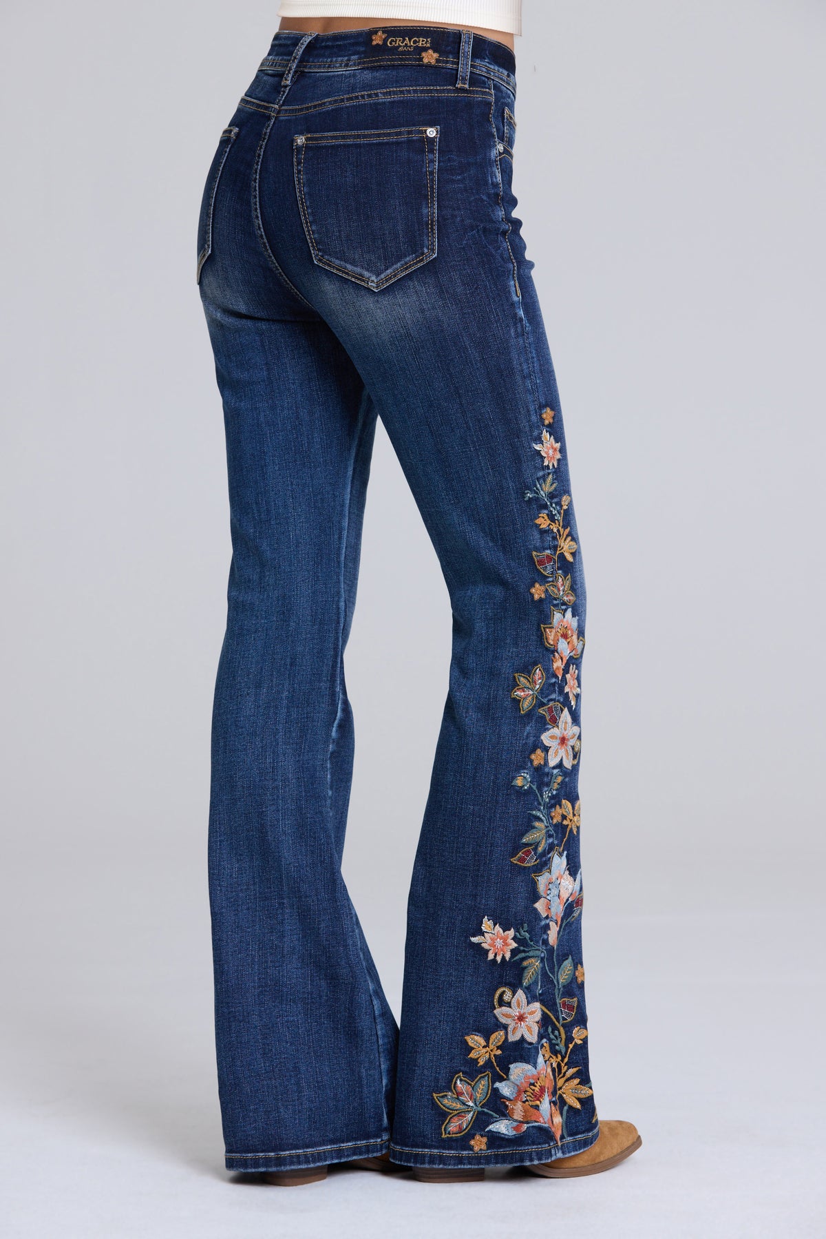 Women's dark blue denim flare jeans with colorful floral embroidery down the outer leg, shown from a rear three-quarter angle on a model.