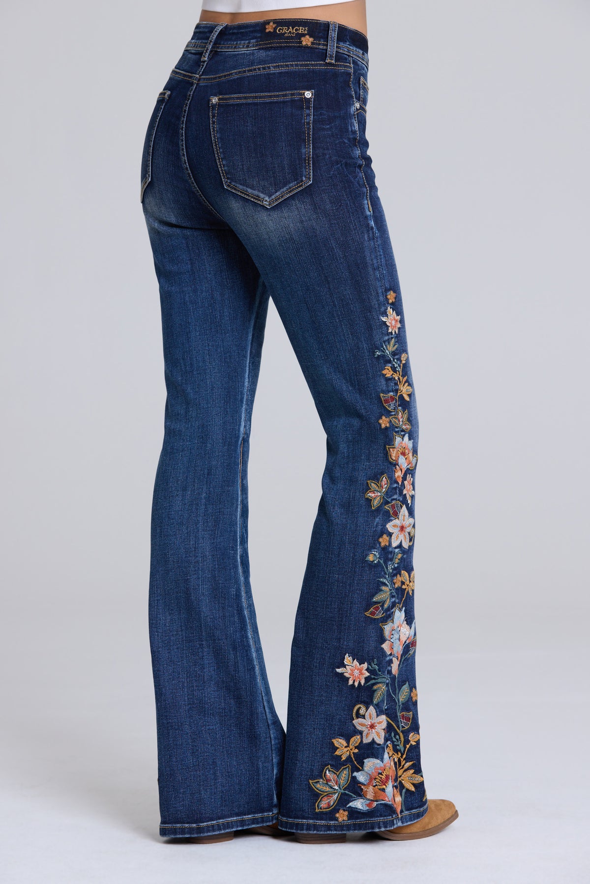 Blue jeans with a high-rise, flare silhouette, featuring detailed floral embroidery down the outer leg on the right side. The jeans are shown from a rear three-quarter angle on a model, set against a plain light gray studio background.