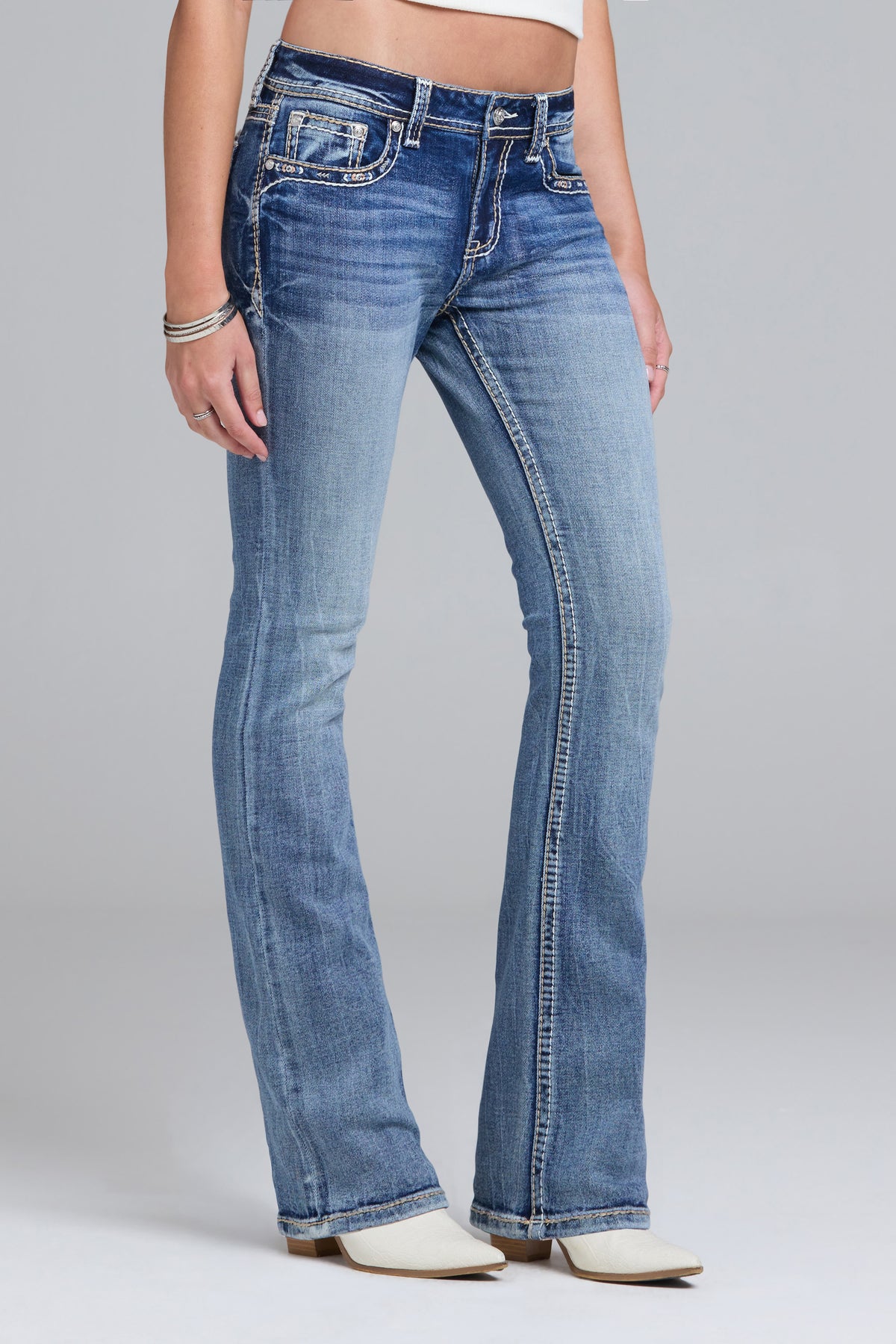 A model wearing a pair of medium-wash blue bootcut jeans with contrast stitching, paired with white heeled boots against a light gray studio background.