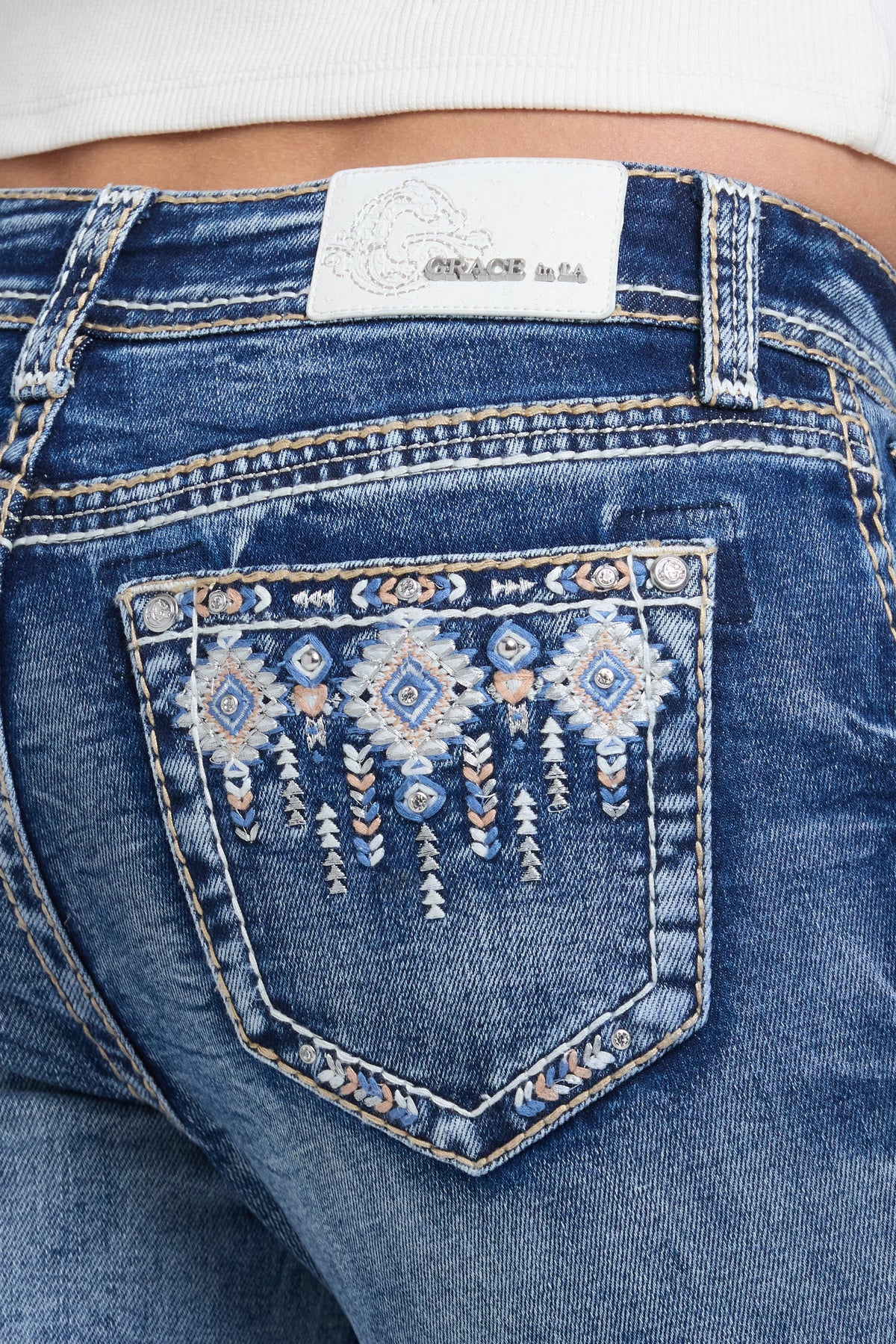 Close-up of the back pocket of blue denim jeans with decorative embroidered detailing in white, blue, and orange, showing a white brand label at the waistband.