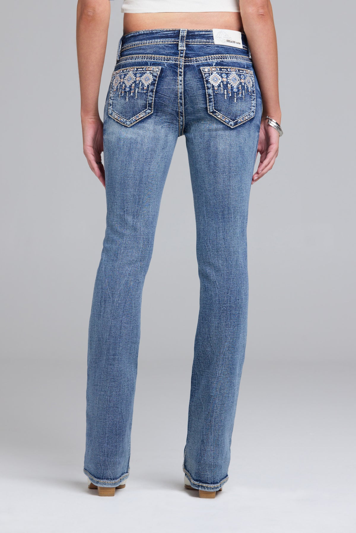 Rear view of a model wearing blue denim jeans with decorative embroidered detailing on the back pockets, set against a plain light gray studio background.