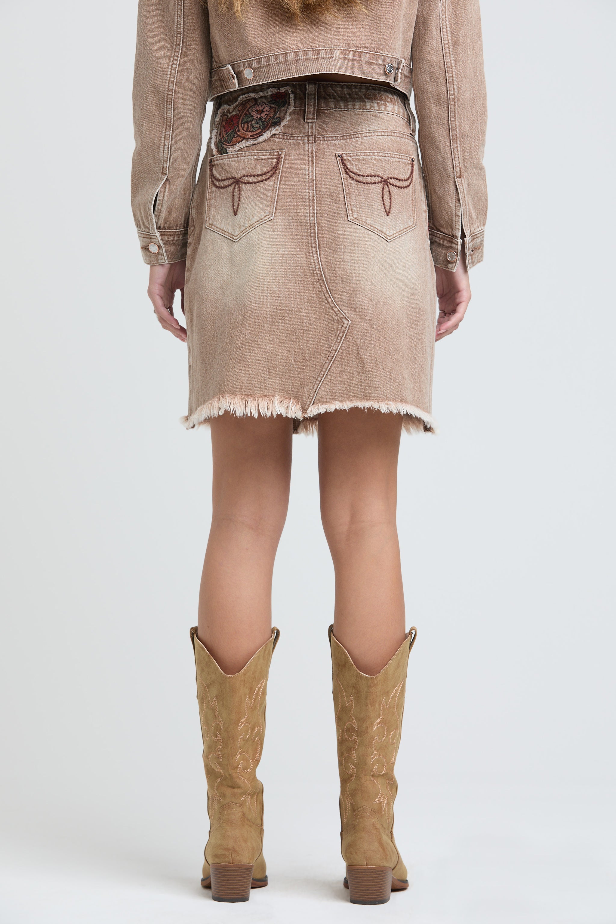 Grace in LA womens tan denim mini skirt featuring longhorn-style embroidery on the back pockets and a frayed hem, styled with tall tan cowgirl boots.