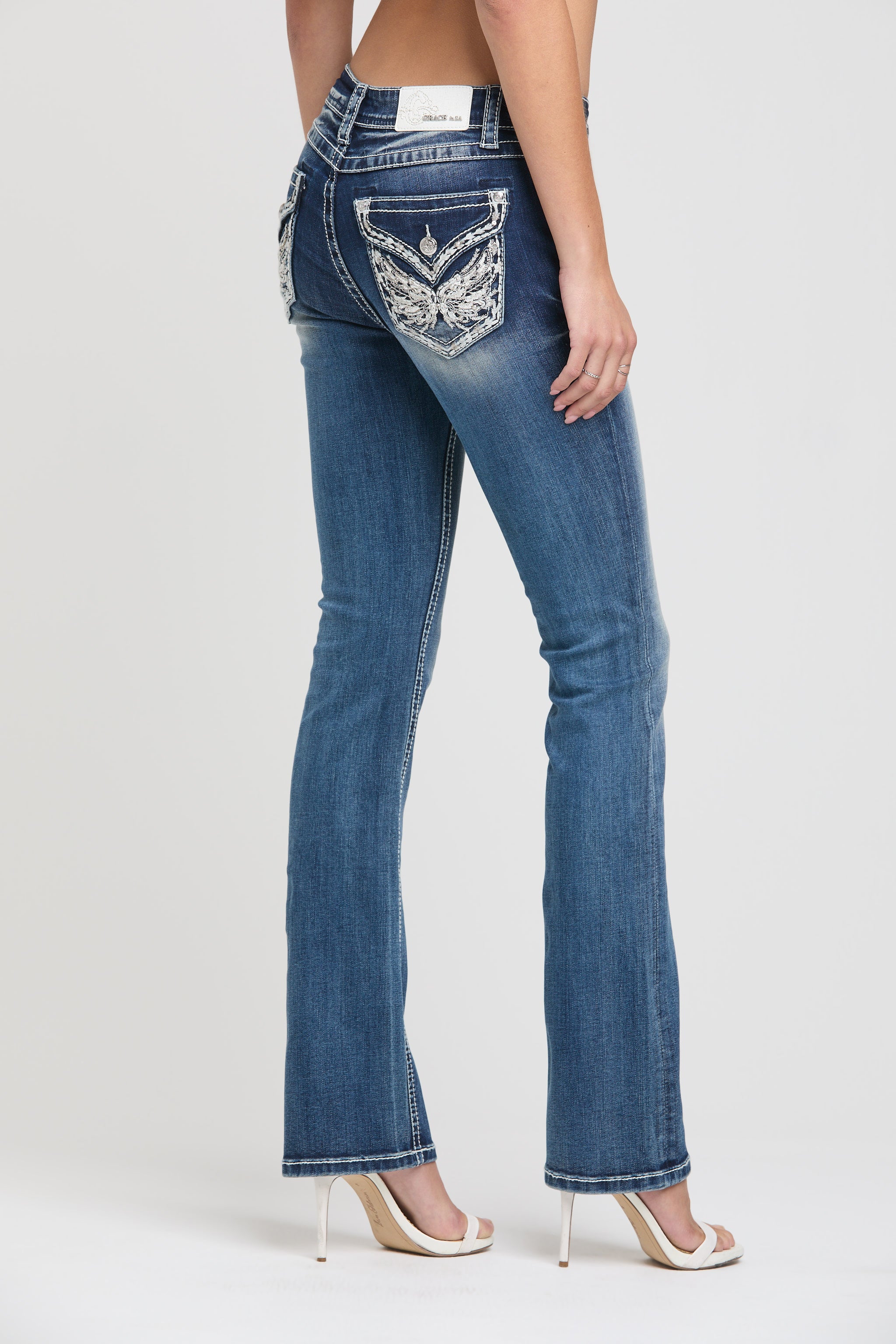 Grace in LA womens bootcut jeans in a dark featuring a rear-facing view with white and silver floral wing embroidery and crystal studs on the back pocket.