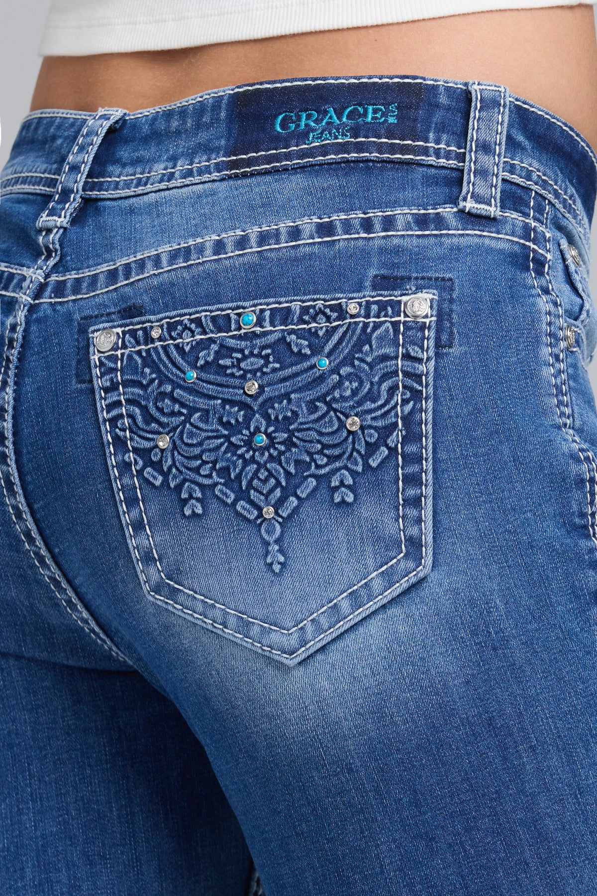 Close-up back view of blue denim jeans with a heavily embellished embroidered pocket and rhinestone detailing, branded GRACE IN LA JEANS on the waistband.