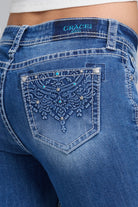 Close-up back view of blue denim jeans with a heavily embellished embroidered pocket and rhinestone detailing, branded GRACE IN LA JEANS on the waistband.