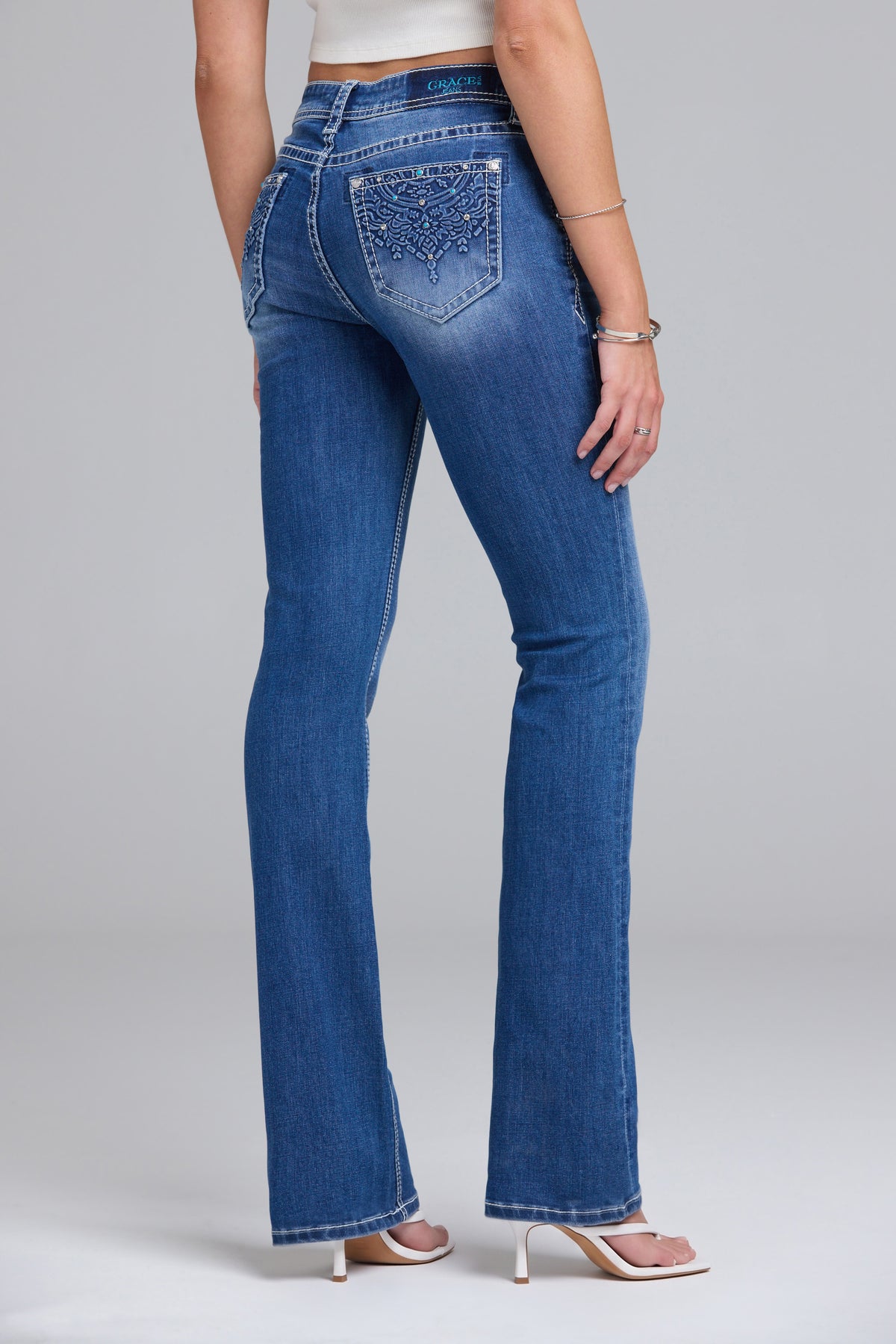 A model from the back wearing blue denim bootcut jeans with silver rhinestone embellishments on the back pocket, paired with a white crop top and white heeled sandals, set against a light gray studio background.