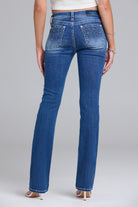 Rear view of a model wearing blue denim jeans with decorative silver rhinestone embellishments on the back pockets, paired with a white crop top and beige high heels against a light gray studio background.