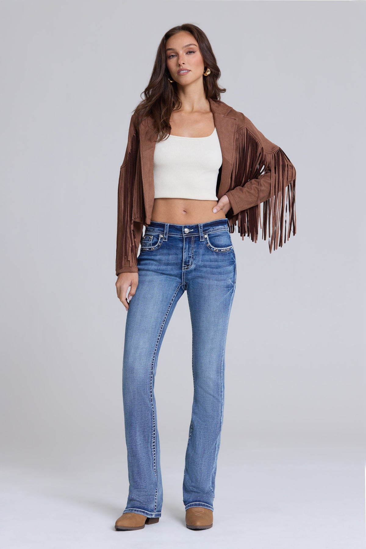 A woman modeling a brown suede fringe jacket, white crop top, and blue jeans against a light gray studio background.