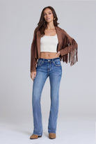 A woman modeling a brown suede fringe jacket, white crop top, and blue jeans against a light gray studio background.