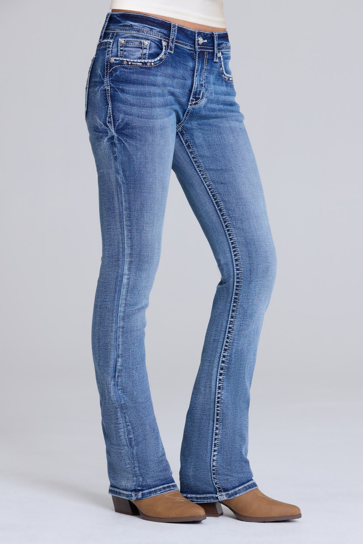 A person wearing medium-wash blue bootcut jeans with brown heeled boots, shown from the waist down against a light gray studio background.