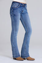 A person wearing medium-wash blue bootcut jeans with brown heeled boots, shown from the waist down against a light gray studio background.