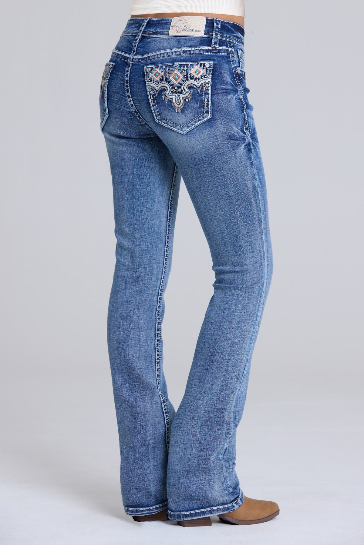 Rusty blue bootcut jeans with intricate white and gold embroidered detailing on the back pocket, shown on a model from a rear three-quarter view against a light gray studio background.