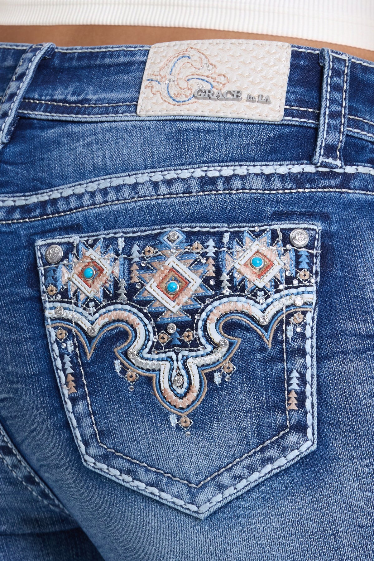 Close-up of the back pocket of blue denim jeans with ornate western embroidery and turquoise rhinestone embellishments. The brand label 'Grace in LA' is visible at the waistband.
