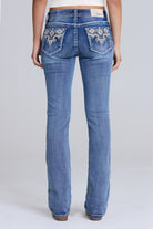 Rear view of a model wearing blue denim jeans with ornate silver and white embroidered detailing on the back pockets, set against a plain light gray studio background.
