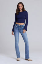 A woman modeling light-wash blue jeans with a navy blue long-sleeve crop top and beige ankle boots, standing against a light gray studio background.