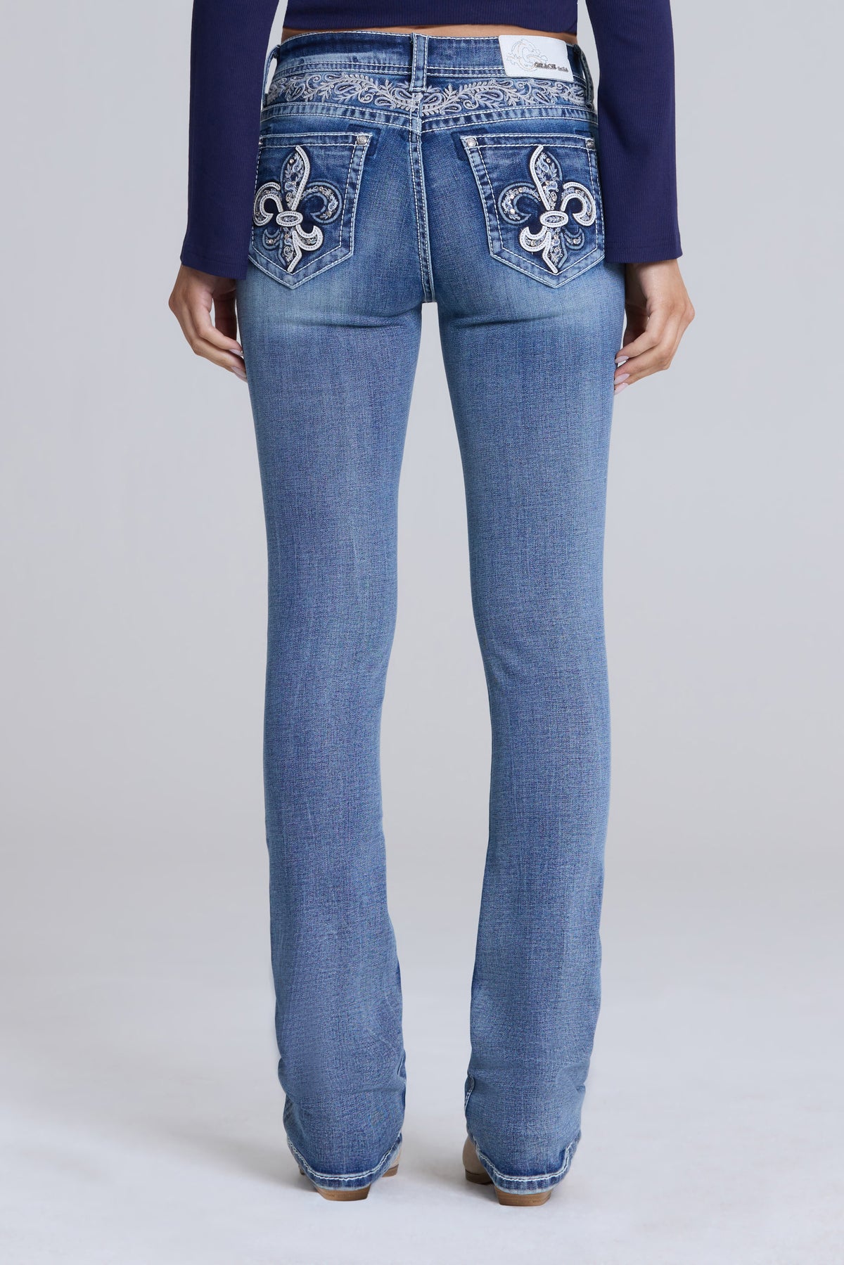 Rear view of a model wearing blue denim jeans with ornate silver fleur-de-lis embroidery on the back pockets, paired with a dark navy long-sleeve top against a light gray studio background.