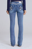 Rear view of a model wearing blue denim jeans with ornate silver fleur-de-lis embroidery on the back pockets, paired with a dark navy long-sleeve top against a light gray studio background.