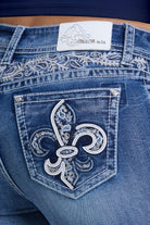 Close-up of the back pocket of blue denim jeans featuring a large embroidered fleur-de-lis motif in silver and white on the pocket, with additional silver embroidery across the waistband and a white brand patch reading 'Grace in LA' visible at the top.