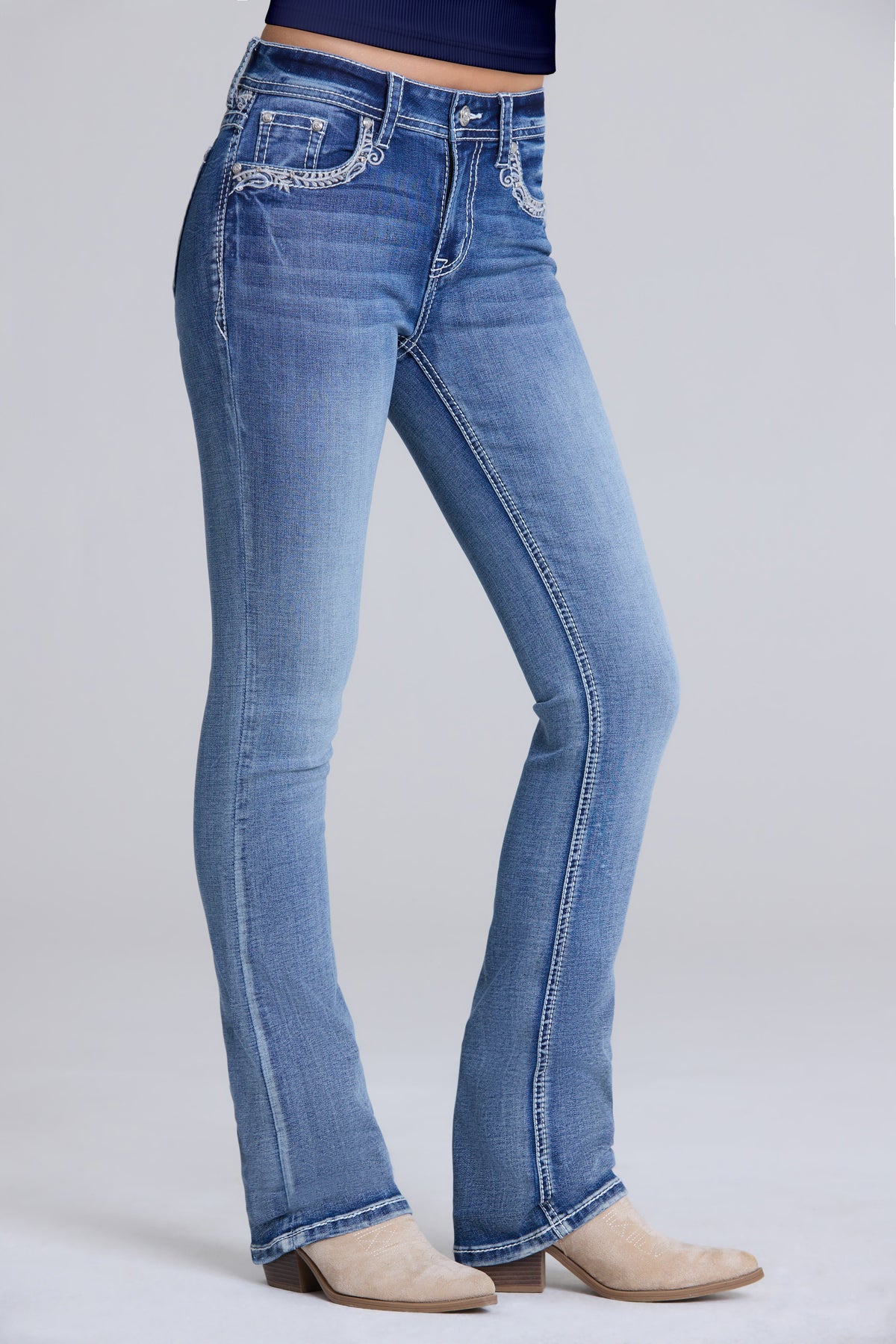 A person wearing light blue denim bootcut jeans with decorative silver embroidery on the front pockets, paired with beige heeled boots against a light gray studio background.