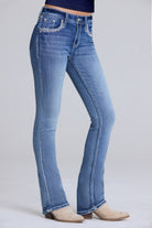 A person wearing light blue denim bootcut jeans with decorative silver embroidery on the front pockets, paired with beige heeled boots against a light gray studio background.