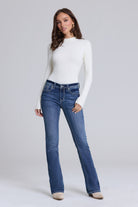 A woman modeling a pair of blue jeans with a white long-sleeve mock-neck top, standing against a light gray studio background.