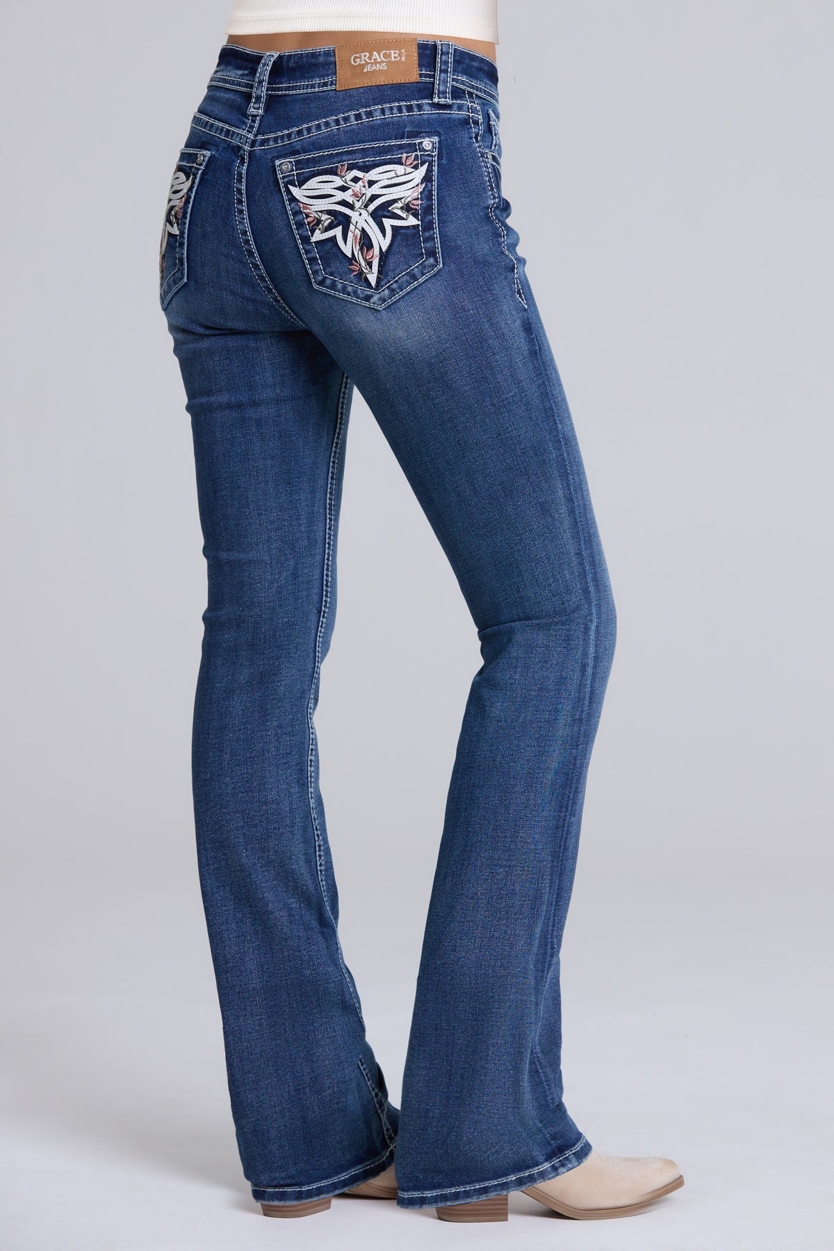A person wearing blue denim bootcut jeans with a large embroidered winged patch on the back pocket, shown from the back against a light gray studio background.