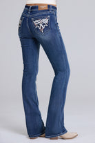 A person wearing blue denim bootcut jeans with a large embroidered winged patch on the back pocket, shown from the back against a light gray studio background.
