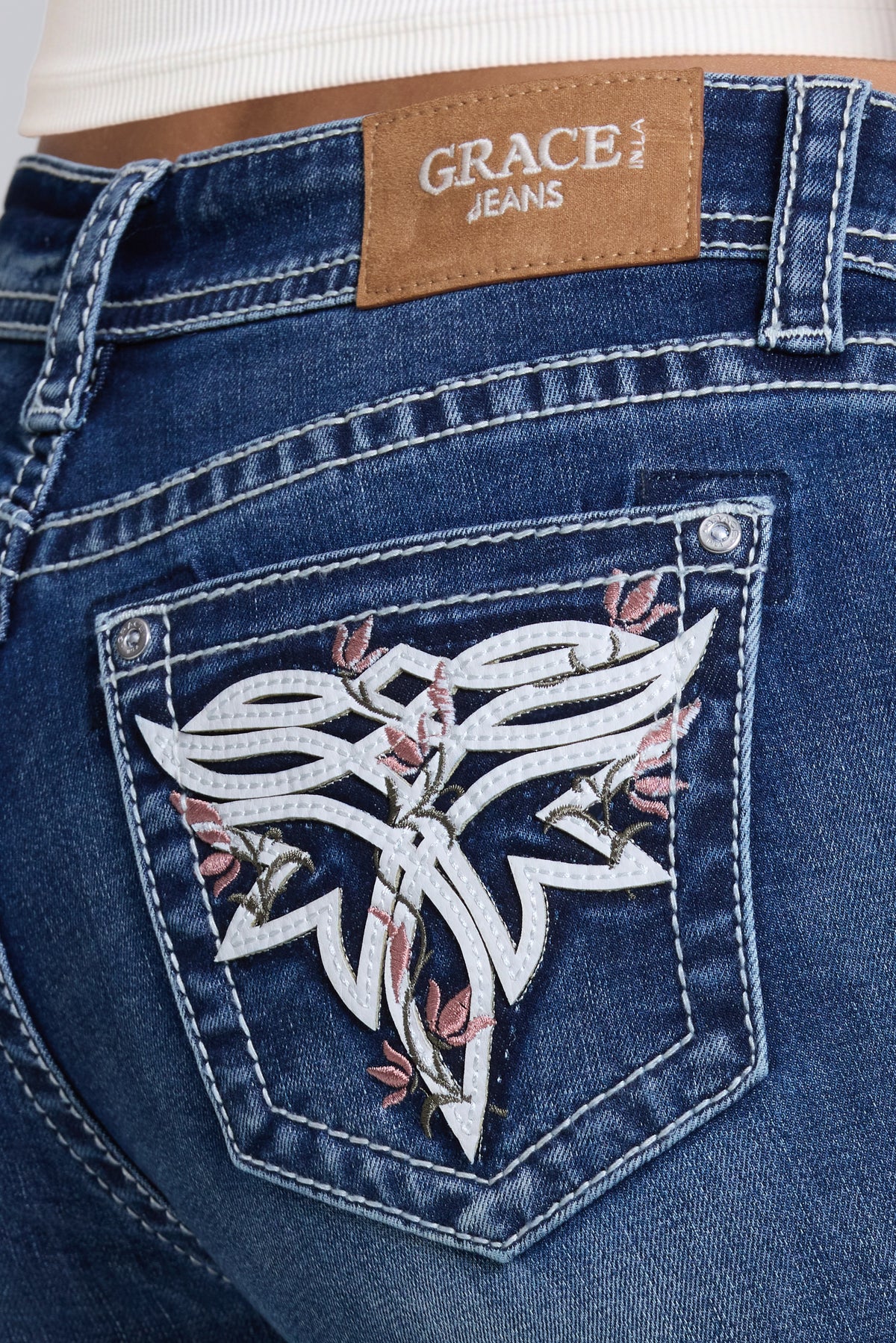Close-up of the back pocket of blue denim jeans with a brown patch reading Grace in LA Jeans and a large white embroidered toe bug graphic with pink floral accents.
