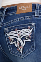 Close-up of the back pocket of blue denim jeans with a brown patch reading Grace in LA Jeans and a large white embroidered toe bug graphic with pink floral accents.