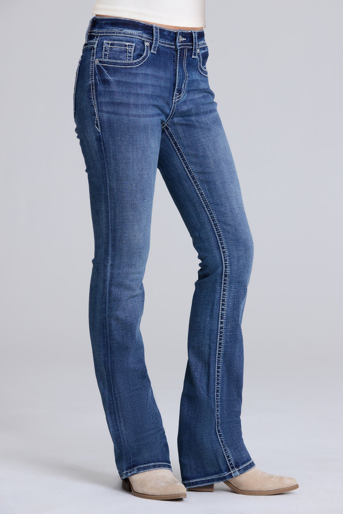 A person wearing blue denim bootcut jeans with a white top and beige shoes, shown from the waist down against a light gray studio background.