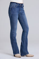 A person wearing blue denim bootcut jeans with a white top and beige shoes, shown from the waist down against a light gray studio background.