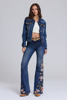 A model stands against a light gray studio background wearing a dark blue denim jacket and matching flare jeans with floral embroidery down the legs, styled with a white crop top and tan boots.