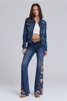 A model stands against a light gray studio background wearing a dark blue denim jacket and matching high-waisted flare jeans with floral embroidery down the legs, styled with a white crop top, tan belt, and tan boots.