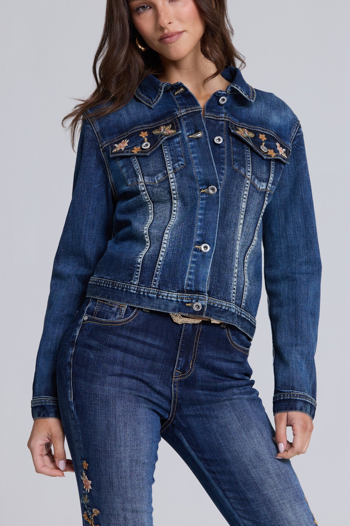 A woman wearing a cropped dark blue denim jacket with floral embroidery on the chest pockets, paired with matching blue jeans, against a light gray studio background.