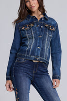 A woman wearing a cropped dark blue denim jacket with floral embroidery on the chest pockets, paired with matching blue jeans, against a light gray studio background.