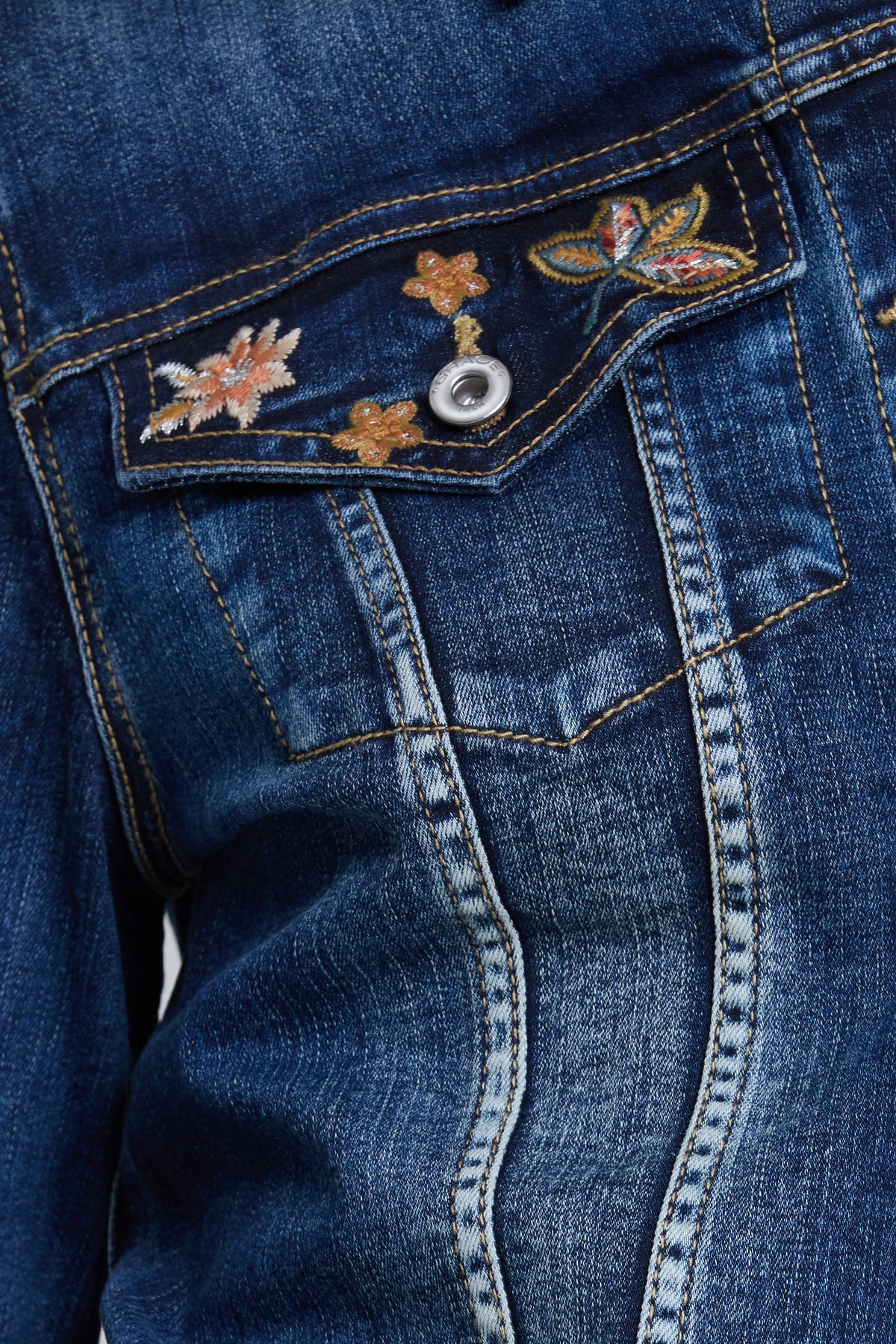 Close-up of a blue denim jacket showing the front pocket with embroidered floral and butterfly details and a silver snap button.