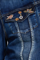 Close-up of a blue denim jacket showing the front pocket with embroidered floral and butterfly details and a silver snap button.