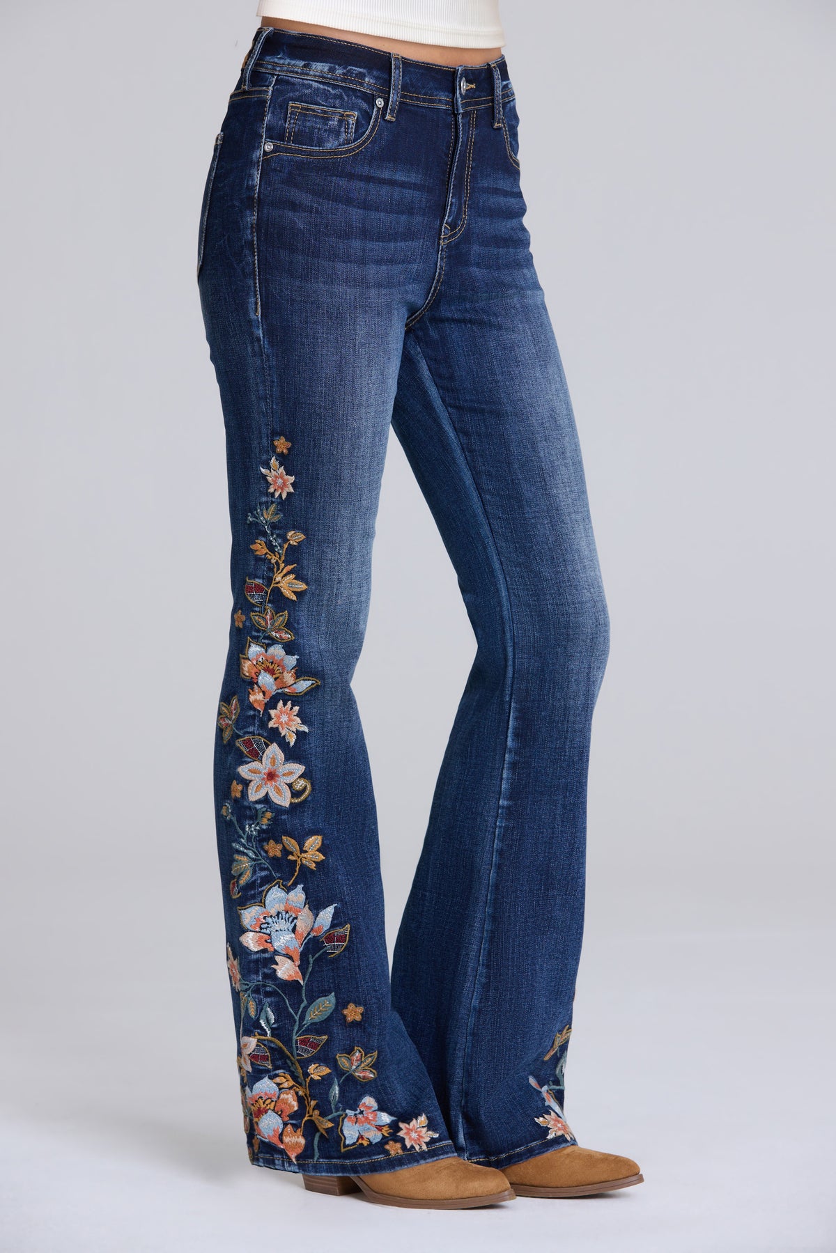 A person wearing blue denim flare jeans with colorful floral embroidery down the front of each leg, paired with tan boots against a light gray studio background.