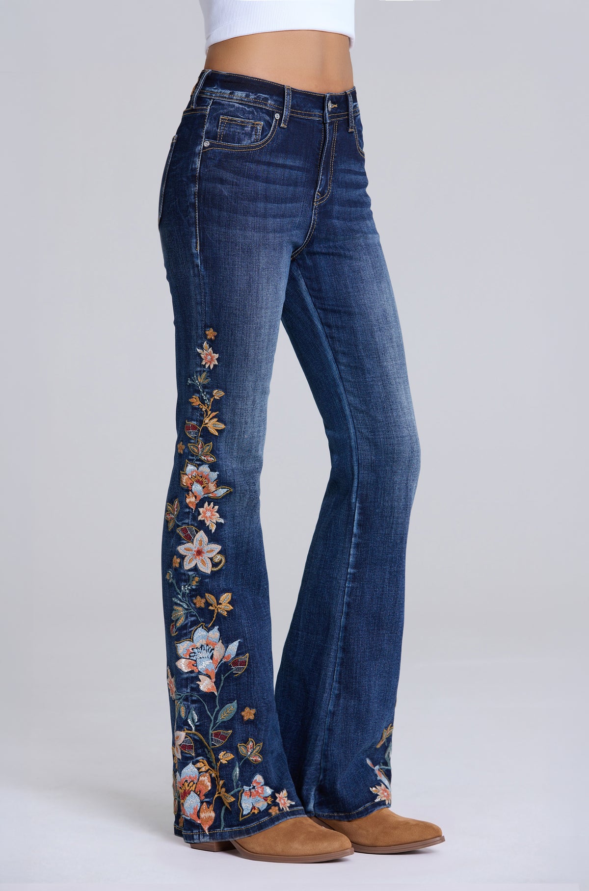 A model is wearing a pair of dark blue denim flare jeans with colorful floral embroidery down the front of the legs, paired with brown suede boots against a light gray studio background.
