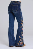 Women's dark blue denim flare jeans with colorful floral embroidery down the outer leg, shown from a rear three-quarter angle on a model.