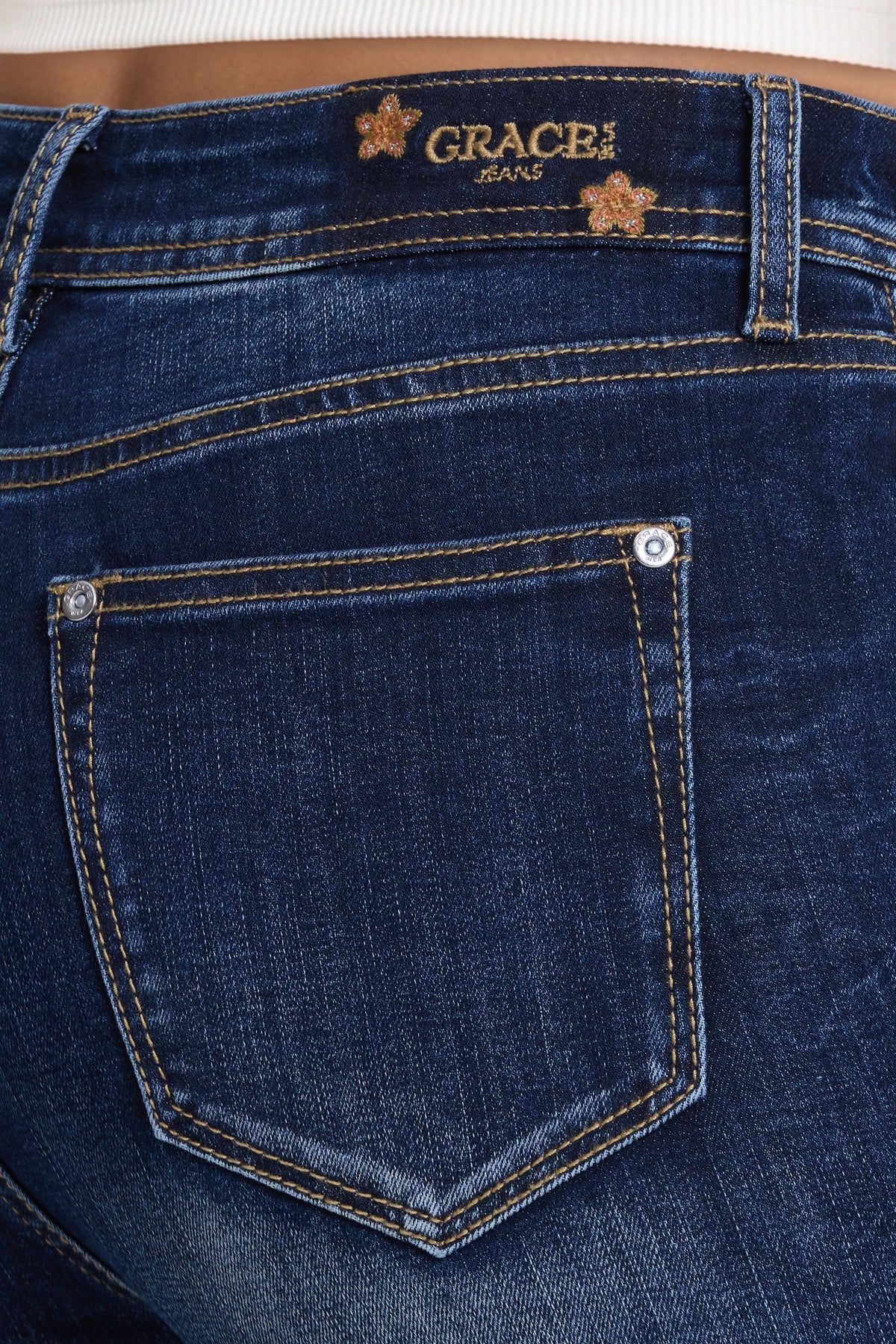 Close-up of the back waistband and pocket of dark blue denim jeans, showing the Grace in LA Jeans logo patch and contrast stitching.